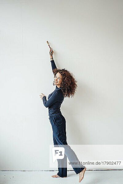 Smiling young woman with hand raised using paintbrush standing on white wall