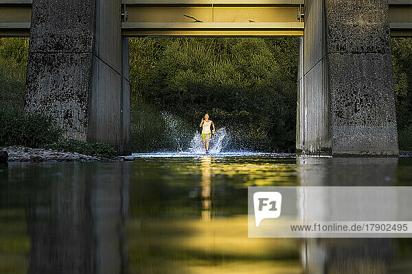 Active young woman running through river water under bridge