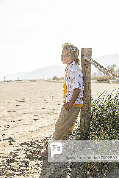 Smiling boy with hand in pocket leaning on wooden pole at beach
