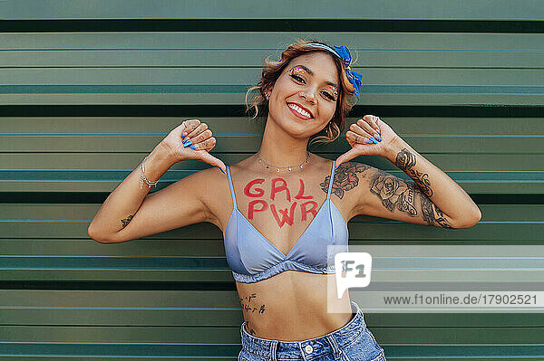 Glückliche junge Frau zeigt Girl-Power-Text auf der Brust vor grüner Wand