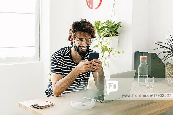 Happy freelancer using mobile phone sitting by laptop at home office