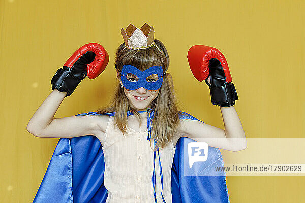 Girl in cape wearing mask and boxing gloves against yellow background