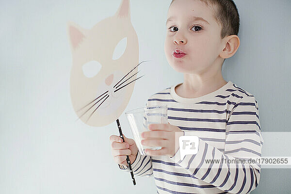 Boy drinking glass of milk and holding mask by wall