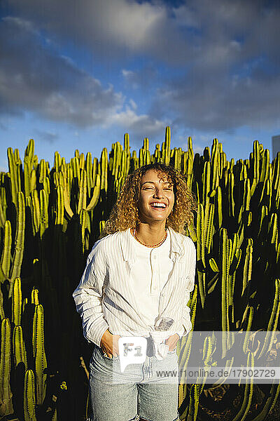 Happy young woman with hands in pockets standing in front of cactus
