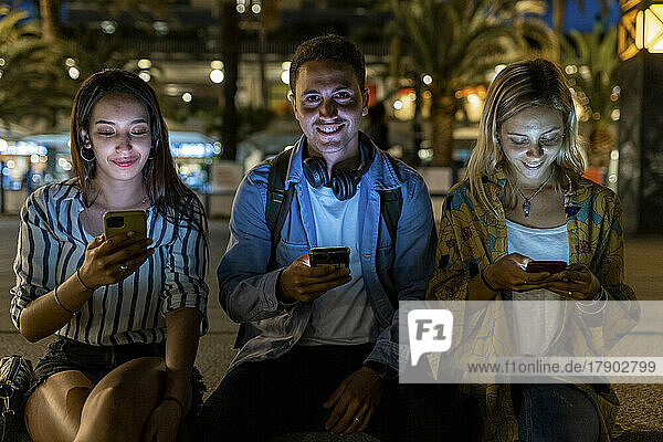 Smiling young man sitting amidst women using smart phone at night