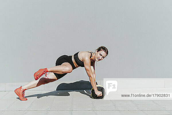 Young athlete with medicine ball exercising on footpath by gray wall