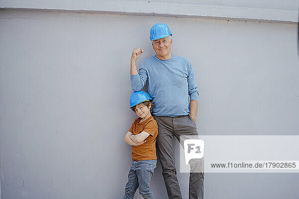 Confident senior man and boy with hardhat in front of wall