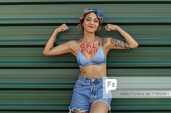 Woman with girl power text on chest showing biceps in front of green metal wall