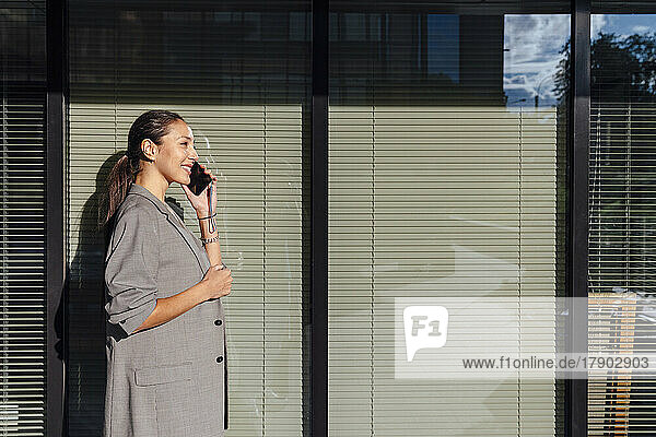 Smiling businesswoman talking on smart phone in front of glass wall
