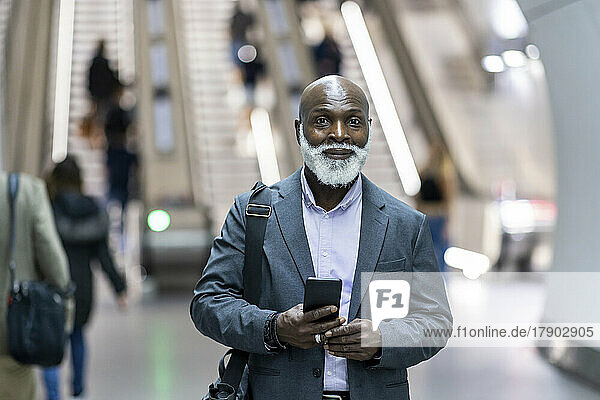 Lächelnder älterer Geschäftsmann mit Laptoptasche und Mobiltelefon am Bahnhof