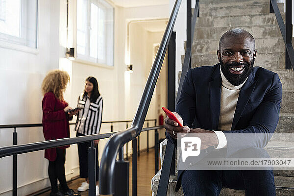 Smiling businessman with smartphone sitting on stairs colleagues talking in background