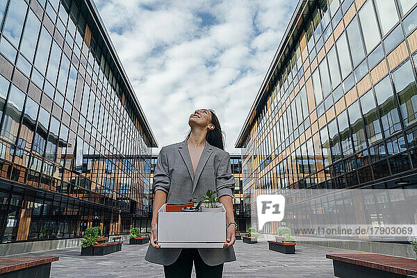 Smiling businesswoman carrying box in front of office building under cloudy sky