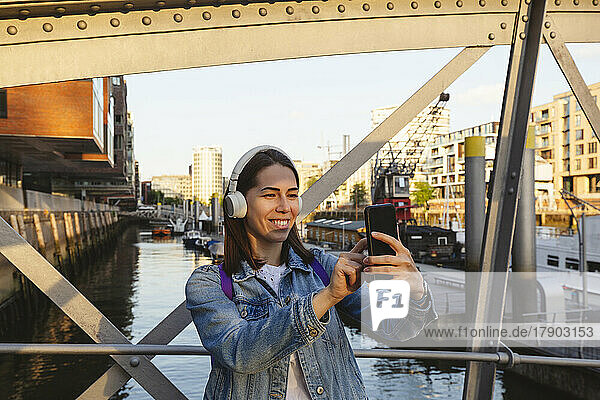 Happy woman wearing wireless headphones taking selfie through smart phone on bridge  Hafencity  Hamburg  Germany