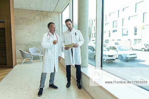 Doctors in lab coats discussing over medical record standing by window at hospital