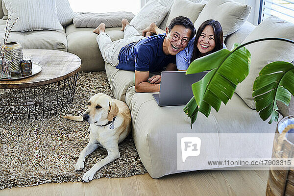 Cheerful couple with laptop lying on sofa in living room at home