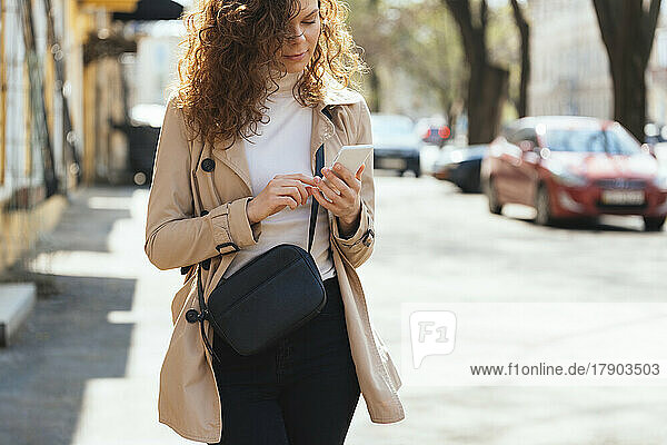 Woman with shoulder bag using mobile phone at sidewalk on sunny day