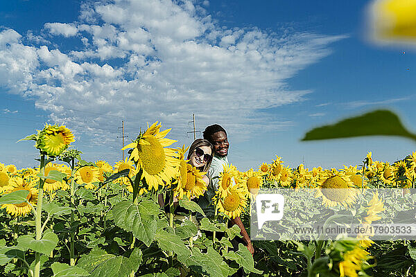 Smiling woman and friend standing back to back in sunflower field on sunny day