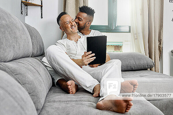 Young man biting ear of boyfriend sitting on sofa at home