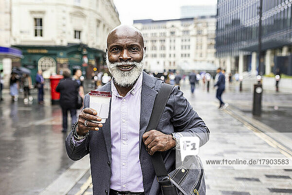 Lächelnder älterer Geschäftsmann mit Laptoptasche beim Kaffeetrinken auf dem Fußweg