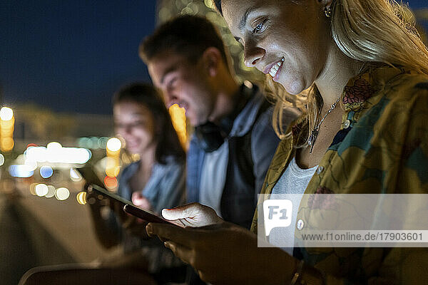 Smiling young woman using mobile phone sitting by friends at night