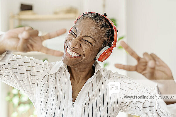 Cheerful woman wearing wireless headphones showing peace sign at home