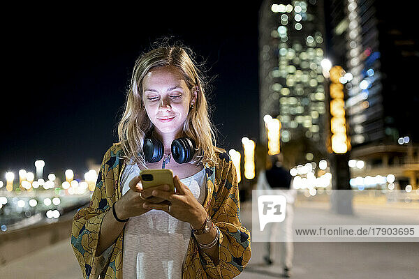 Blonde Frau mit kabellosen Kopfhörern nutzt nachts ihr Smartphone