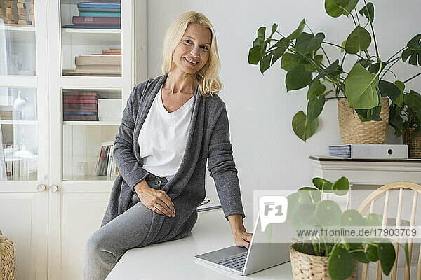 Smiling freelancer with laptop sitting on desk in home office