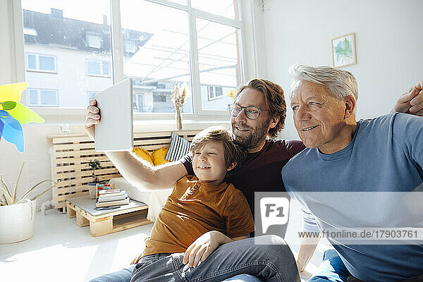 Smiling man taking selfie with son and father through tablet PC at home