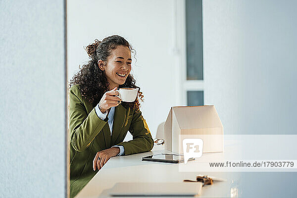 Glückliche Geschäftsfrau mit Kaffeetasse und Modellhaus auf dem Schreibtisch im Büro