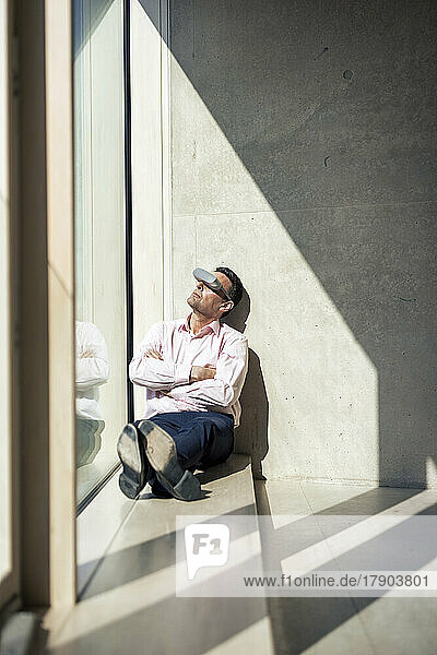 Mature businessman watching through futuristic glasses sitting on window sill in office