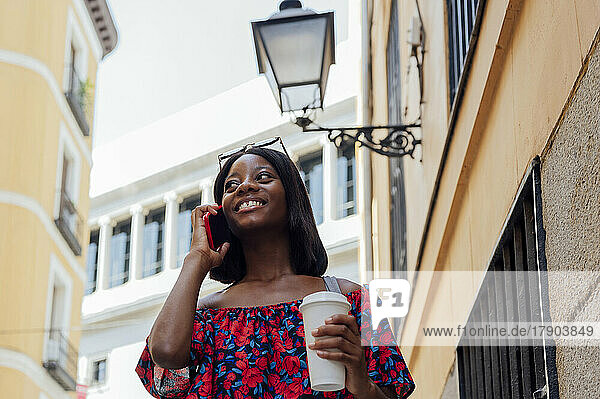 Smiling woman holding disposable coffee cup talking on smart phone by building