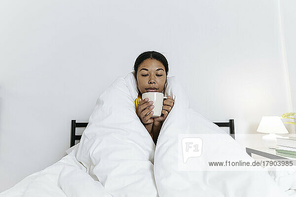 Woman sitting on the bed wrapped in a blanket drinking tea