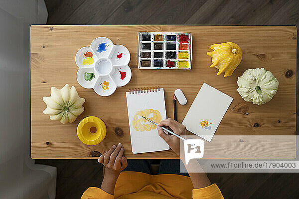 Mädchen mit Aquarellfarben malt Jack O'Lantern auf einem Skizzenblock am Tisch