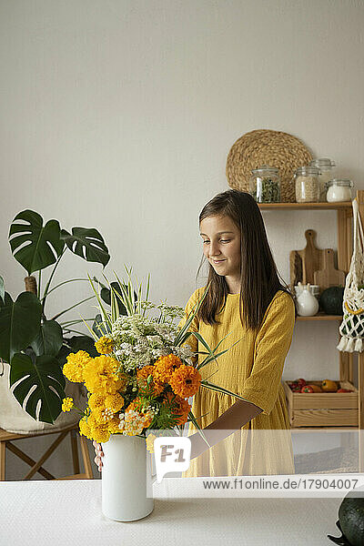 Smiling girl holding flower vase standing by table in kitchen at home