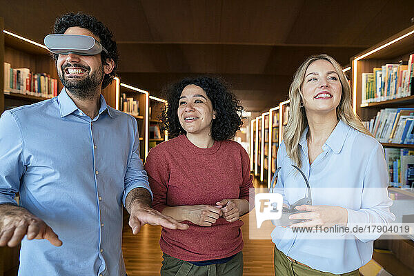 Smiling man watching through VR glasses by colleagues in library
