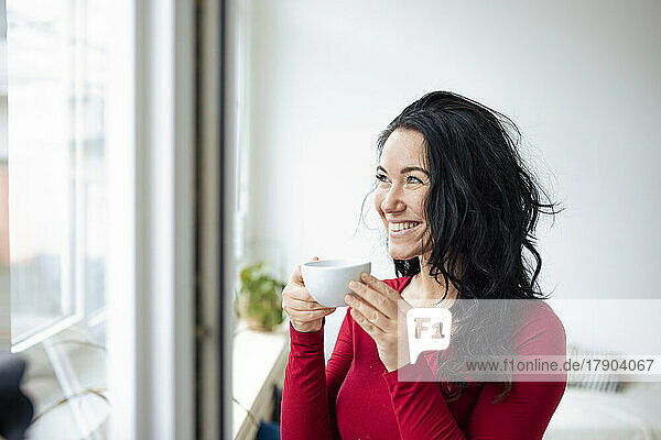 Glückliche Frau mit Kaffeetasse und Blick durch das Fenster