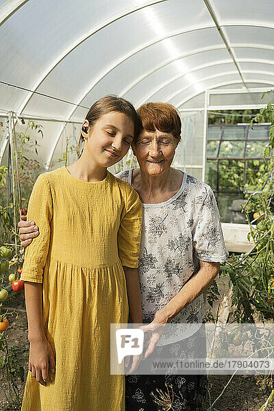Lächelnde ältere Frau und Enkelin mit geschlossenen Augen stehen zusammen im Gewächshaus