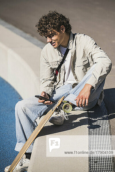 Smiling man with skateboard using smart phone sitting at edge of track and field