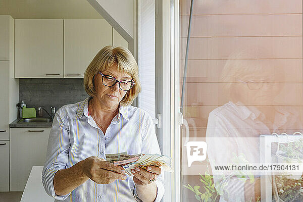 Senior woman counting paper currency standing by glass window at home