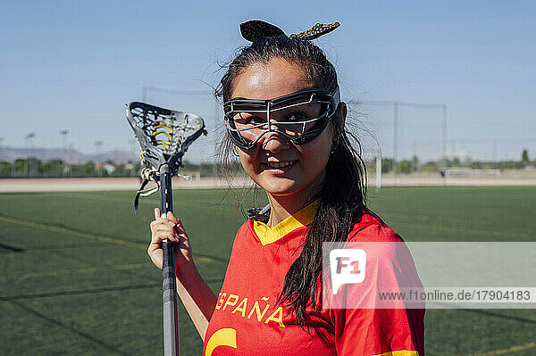 Lächelnder Spieler mit Schutzbrille  der an einem sonnigen Tag einen Lacrosse-Stick hält