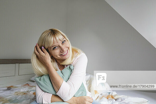 Glückliche blonde Frau mit Kissen sitzt zu Hause im Bett