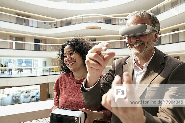 Smiling businessman with VR glasses gesturing by colleague in corridor