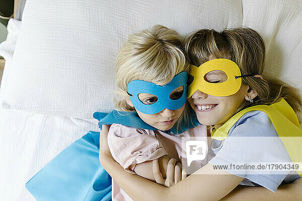 Lächelndes Mädchen im Superwoman-Kostüm umarmt Schwester zu Hause im Bett