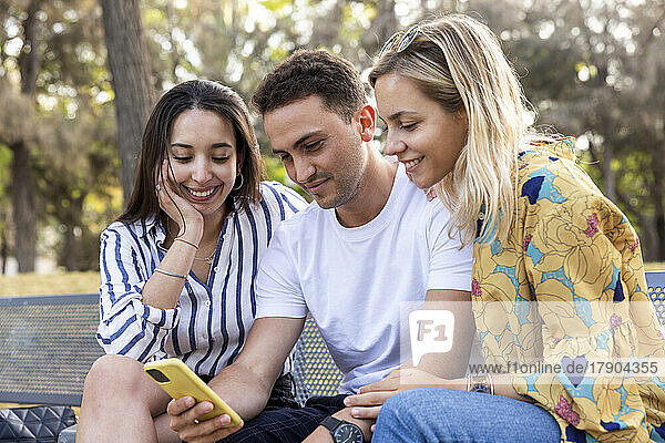 Lächelnder junger Mann  der im Park sein Smartphone mit Freunden teilt
