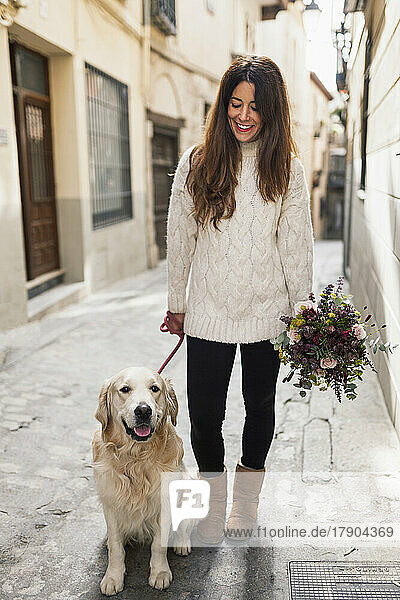 Lächelnde Frau mit Hund hält Blumenstrauß in der Gasse