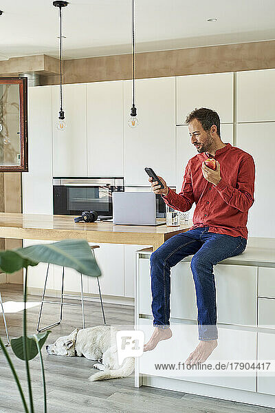 Smiling man using smart phone and eating apple at home