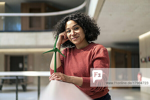 Smiling businesswoman with head in hand holding wind turbine on railing in office corridor