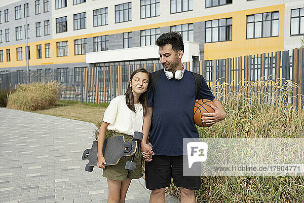 Lächelndes Mädchen mit geschlossenen Augen  das die Hand seines Vaters hält  der Basketball trägt  vor dem Gebäude