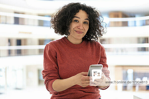 Lächelnde Geschäftsfrau mit lockigem Haar hält Smartphone im Büro