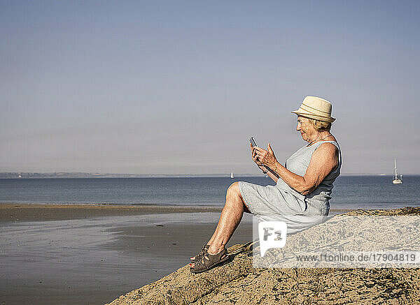 Senior woman using tablet PC sitting on rock at beach